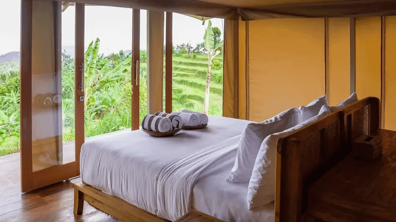 Habitación de hotel con cama blanca y toallas enrolladas, rodeada de ventanales con vistas a una vegetación tropical.