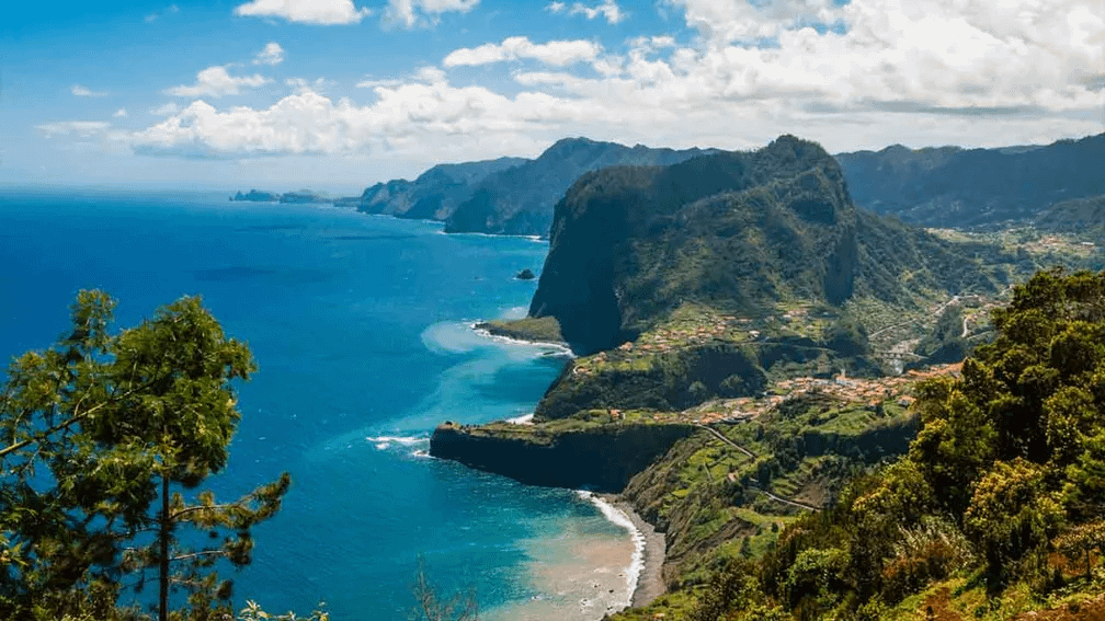 Vista panorámica de la costa de Madeira, con acantilados verdes, mar azul y una pequeña playa; el paisaje señala el destino del próximo viaje.