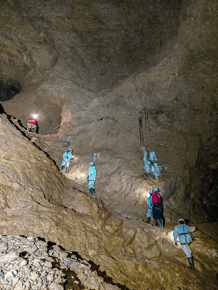 Varias personas con cascos y arneses avanzan por una vía ferrata sobre una pared rocosa dentro de una enorme cueva oscura.
